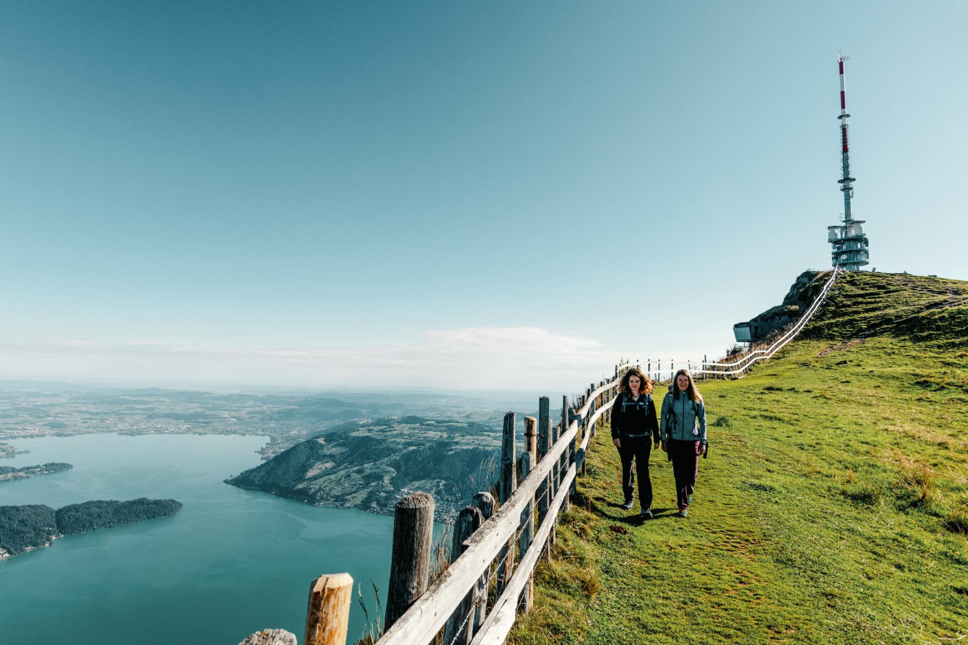 Tell-Trail Etappe 3: Zwei Wanderinnen auf der Rigi mit der Gipfelantenne und dem Zugersee im Hintergrund.